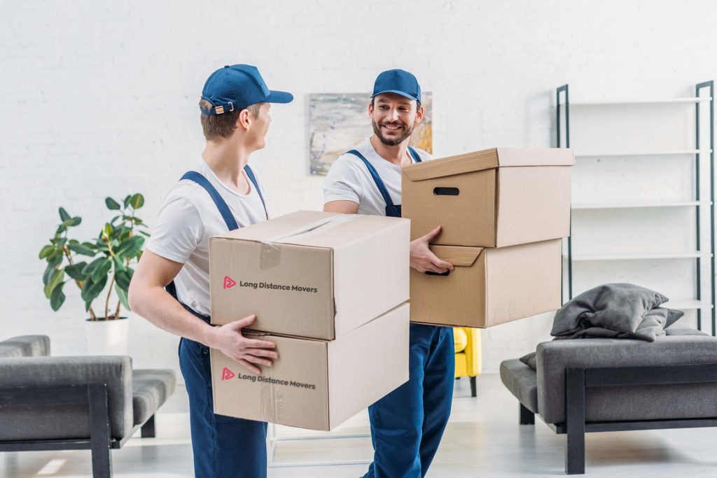 two movers in uniform looking at each other while carrying cardboard boxes in apartment