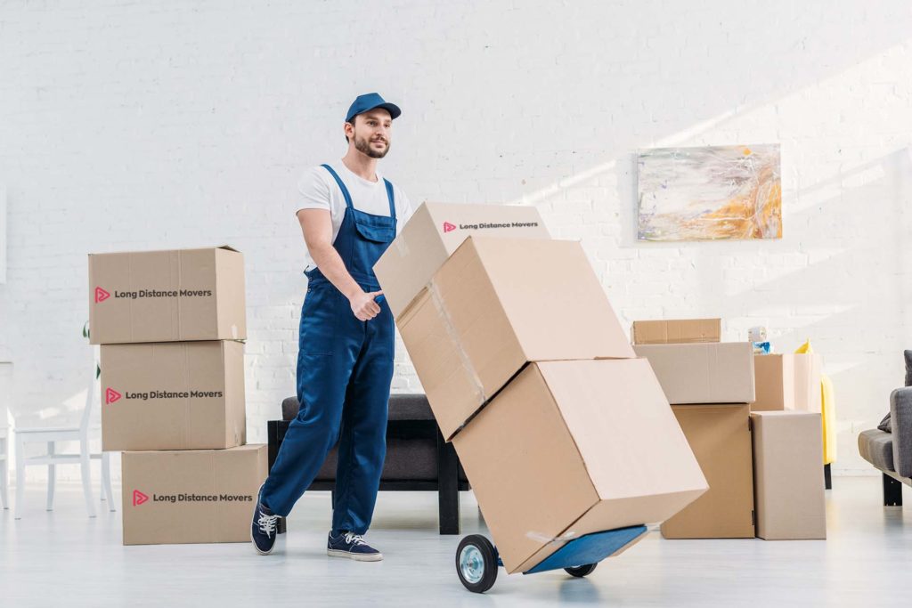 mover in uniform transporting cardboard boxes on hand truck in apartment