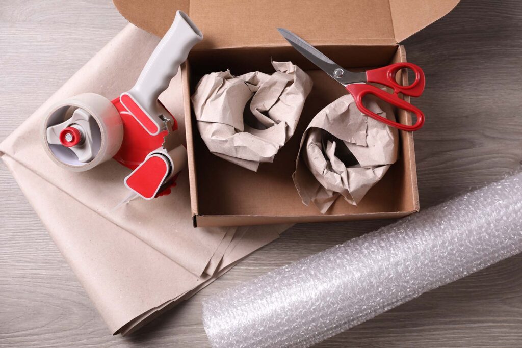 Open box with wrapped items, adhesive tape, scissors, paper and bubble wrap on wooden table, flat lay